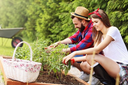 A young man and woman are gardening together outdoors in a well-maintained garden near Westminster, with lush green trees and shrubs in the background. They are kneeling on the soil beside a flower bed, actively tending to the plants. The man, dressed in a red and blue plaid shirt and a straw hat, is using his hands to support or prune the plants. The woman, wearing a white t-shirt and a red headband, is also engaged in planting or trimming, with a gentle smile on her face. To the left, a white wicker basket filled with gardening tools and plant debris is placed on the edge of the flower bed, indicating ongoing gardening activity. The garden surface features a mixture of rich, dark soil and neatly edged planting borders, with patches of grass nearby. The overall scene suggests a pleasant day of outdoor gardening, reflecting a landscaped outdoor space likely maintained by local gardening services, such as those offered by Landscapers Westminster, operating in the SW1 postcode area, with natural lighting indicating a partly cloudy day.