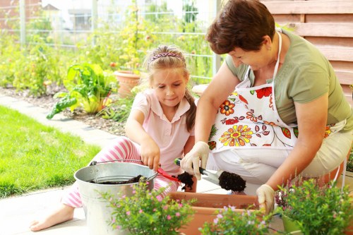 In a well-maintained garden, a woman and a young girl are engaged in planting or gardening activities on a sunny day. The garden features a lush green lawn in the foreground, bordered by flower beds with vibrant pink and purple flowering plants. Behind them, there is a wooden garden shed or fence, providing a backdrop of natural wood tones. The background includes a variety of garden elements such as shrubs, small trees, and a trellis with climbing plants, contributing to a layered landscape design. The woman is wearing a light green top and a colorful apron, while the girl has long hair tied back and is dressed in a pale pink top with striped trousers. They are both actively working with gardening tools, such as small trowels, over a rectangular flower bed or planter box, surrounded by gardening pots and a metal watering can. The scene captures a warm, sunny weather condition, with dappled sunlight filtering through the garden foliage, creating a inviting atmosphere indicative of outdoor gardening service environments in Westminster, supporting lush, healthy outdoor spaces maintained by local landscapers.