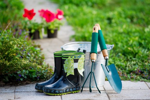 A pair of dark rubber gardening boots with a decorative pattern along the sole, placed on a paved stone path in a garden. Next to the boots, there is a green and black striped garden glove hanging from a small metal clip, and a pair of gardening shears with wooden handles resting on top of a blue trowel. Behind these tools, a white plastic bucket is visible, likely used for planting or watering. In the background, there are vibrant pink flowers in pots along the garden border, with lush green foliage and grass extending further back. The scene suggests a well-maintained outdoor space suitable for gardening or lawn care in Westminster, with a bright, clear weather condition typical of a spring or summer day, highlighting a setup for gardening tasks managed by landscapers Westminster.