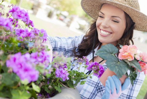 A woman in a wide-brimmed straw hat and light blue gardening gloves is tending to a vibrant flower bed in a residential garden, with a variety of colourful flowering plants including pink, purple, and peach blooms. The garden features a well-maintained lawn in the foreground, bordered by flower beds with lush green foliage and neatly arranged soil. In the background, there are mature trees and a hedge providing privacy, with soft natural lighting indicating a bright, partly sunny day. The scene is set outdoors in a typical Westminster garden, showcasing a peaceful, landscaped outdoor space suitable for gardening and outdoor maintenance services by landscapers Westminster. The woman is smiling warmly as she carefully tends the plants, emphasizing the care and attention to detail involved in maintaining a visually appealing garden environment.
