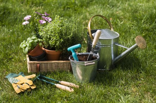 The image depicts a well-maintained garden scene with a vibrant green lawn in the foreground, showing dense, healthy grass. To the left, a wicker basket contains various potted plants, including flowering plants with pink and purple blooms, along with lush green foliage. Nearby, gardening tools such as a pair of yellow gardening gloves, small hand trowels with wooden handles, and a blue-handled transplanter rest on the grass. To the right, there is a galvanized metal watering can with a wooden handle, positioned next to a small metal bucket that holds a white-handled garden hand rake. The backdrop features additional greenery, suggesting a lush outdoor area suitable for garden care and landscaping activities. The natural lighting indicates a bright, possibly sunny day, enhancing the vibrant colours and textures of the garden elements. This scene captures tools and plants typically associated with gardening services offered by Landscapers Westminster in Westminster, London, emphasizing garden maintenance and outdoor landscaping tasks in a residential or commercial garden setting.