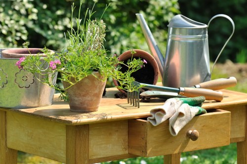 A wooden garden table situated outdoors in a well-maintained garden setting, featuring a variety of gardening tools and containers. The table holds a terracotta pot with lush green herbs and plants, including some with fine, feathery foliage. To the left, there is a metal watering can with a curved handle and spout, alongside a small gardening trowel resting on the table's surface. A pair of gardening gloves, slightly dirty from use, are draped over the edge of the table. In the background, the garden appears vibrant and lush, with blurred greenery and flowering plants, indicating a pleasant weather condition suitable for gardening activities. The scene reflects an active gardening environment, possibly part of a landscaping service in Westminster or a similar area in the UK, emphasizing outdoor maintenance, plant care, and garden planning. The natural daylight illuminates the scene, highlighting the textures of the wooden table and various garden tools, supporting the professional services provided by Landscapers Westminster in creating and maintaining attractive outdoor spaces.