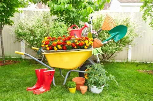 A garden scene featuring a yellow wheelbarrow filled with vibrant red, orange, and yellow flowers positioned on a well-maintained green lawn. Surrounding the wheelbarrow are gardening tools, including a pair of red rubber boots, a red watering can, and various small terracotta pots. In the background, there are lush bushes and a wooden fence, with trees providing partial shade, indicating an outdoor space in Westminster. The overall setting suggests active gardening or planting, with natural light highlighting the fresh and healthy appearance of the plants and outdoor equipment, embodying professional gardening and landscaping care suitable for a residential garden in the area.