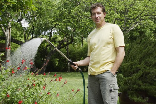 A man in a light yellow t-shirt is standing in a well-maintained garden, watering a flower bed with a garden hose. The garden features a lush, green lawn with dense grass, bordered by a variety of blooming red flowers and leafy plants. Behind him, there are mature trees with green foliage providing shade, alongside neatly trimmed hedges and shrubs, creating a structured landscape. The soil around the flower bed appears healthy and moist, while the paving and decking areas are clean and well-kept. The scene is set outdoors on a bright, overcast day, with natural light highlighting the vibrant colors of the plants and the overall tidy appearance of the garden. This setting exemplifies professional gardening and landscaping standards, as provided by Landscapers Westminster, tailored to outdoor spaces in Westminster or nearby areas within the W postcode region, reflecting expertise in garden care and maintenance services.