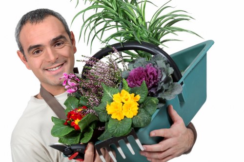 A professionally dressed gardener holds a black plastic tray filled with various potted plants, including purple, yellow, and red flowering varieties, in front of a larger potted green plant with long, arching leaves. The background features part of a landscaped garden with a well-maintained lawn area and neatly arranged flower beds edged with soil. The scene is set outdoors under natural daylight, with a clear sky and soft shadows indicating a bright, calm weather. The garden's layout shows defined grassy spaces and garden borders, contributing to a tidy, inviting outdoor environment suitable for landscape gardening and lawn care services typical for Westminster and surrounding areas. The gardener's friendly expression and the arrangement of plants emphasize professional gardening and planting expertise, supporting landscaping and garden renovation services offered by Landscapers Westminster.