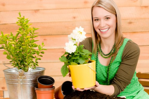The image depicts a woman with blonde hair smiling while sitting outdoors in a garden area, holding a yellow flower pot with a flowering green plant, possibly a geranium. She is dressed casually in a green polka-dot apron over a brown long-sleeve top and is accessorized with layered necklaces. The background includes a wooden fence and a newly planted shrub in a metal bucket, alongside other empty plant pots placed on a wooden surface, suggesting gardening activity. The garden surface shows a mix of soil and paving, with potential grass or lawn areas in the surrounding space. The environment appears well-maintained, natural, and suitable for outdoor gardening services, with visible elements indicating care and planting routines typical of professional landscapers in Westminster or nearby areas. The weather looks clear, with natural light enhancing the vibrant colours of the plants and the woman’s attire, supporting the theme of garden maintenance and landscaping in a typical UK outdoor setting.