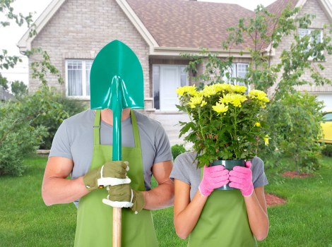 A photograph depicts two gardening enthusiasts standing on a well-maintained front lawn in a suburban neighbourhood. The individual on the left is wearing a light grey t-shirt, a green gardening apron, and tan gardening gloves, holding a small gardening spade with a teal handle, which partially covers their face with a teal plastic hood. The person on the right is dressed in a grey T-shirt, bright pink gardening gloves, and is holding a large bouquet of yellow chrysanthemums in a pink foil wrap, obscuring their face. Behind them, a brick house with white-framed windows, a grey roof, and a small garden with bushes and a tree is visible, along with a neatly edged lawn and flower beds with soil borders. The outdoor scene is lit by natural daylight under slightly overcast skies, reflecting typical spring or summer garden conditions, consistent with professional gardening and landscaping services offered by Landscapers Westminster in Westminster, SW1 area. The overall setting illustrates a clean, landscaped front garden, emphasizing cultivation, planting, and outdoor maintenance.