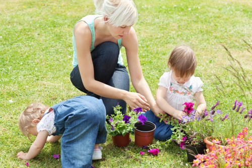 A woman with short platinum blonde hair, dressed in a light blue sleeveless top and dark jeans, is kneeling on a well-maintained grassy garden area in Westminster, engaging with two young children. One child, wearing a white shirt with a floral pattern and blue overalls, is sitting on the grass and examining a flower bed with vibrant purple and pink flowering plants. The other child, also dressed in a similar white top, is crouched lower, reaching towards the ground to explore the soil and plants. Nearby, there are small terracotta pots with young plants, indicating active gardening or planting activities. The garden features a lush, dense lawn with a few patches of slightly uneven grass, bordered by flower beds filled with multi-coloured blossoms. Bright natural light suggests a clear day, emphasizing the vivid colours of the flowers and the greenery. The scene captures a moment of outdoor leisure and gardening, reflecting the professional landscaping services offered by Landscapers Westminster in the local area, contributing to a thriving garden environment in Westminster, SW1 postcode.