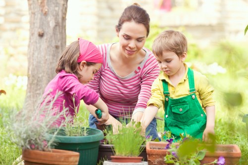 A woman gardening outdoors in a landscaped garden with lush green grass, potted plants, and a flower bed, accompanied by a young girl in a pink headscarf and purple top, and a young boy in a yellow shirt and green apron, all engaged in planting or tending to the plants. The garden features a large tree trunk in the background, natural soil beds, and various gardening containers, with soft sunlight illuminating the scene. The environment appears well-maintained and vibrant, indicative of professional landscape and gardening services in Westminster. The scene illustrates outdoor family gardening activity, highlighting the importance of horticultural care and garden design support provided by local experts like Landscapers Westminster.