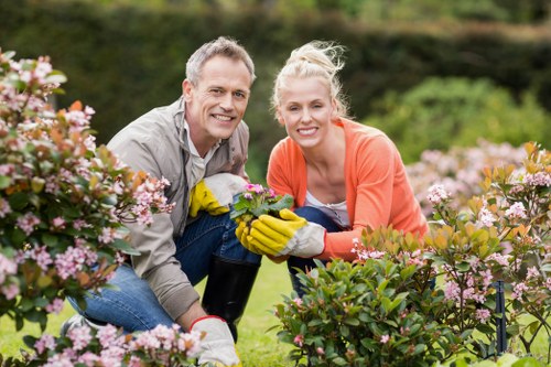 A smiling middle-aged man and woman in a well-maintained garden, kneeling among blooming pink and white flowering shrubs. The man has short grey hair and is wearing a light grey jacket, while the woman has blonde hair tied back and is dressed in an orange top. They are both wearing gardening gloves, and the woman is holding a small potted plant. The garden features lush green grass, neatly edged flower beds, and various bushes, with a background of trees and a clear sky suggesting a bright, sunny day. This outdoor space is part of a landscaped yard maintained by local gardening experts, with a focus on flower bed planting and shrub management, aligning with services offered by Landscapers Westminster in the Westminster area. The scene highlights community gardening activities, emphasizing the importance of outdoor maintenance and plant care, central to professional landscaping and gardening services.